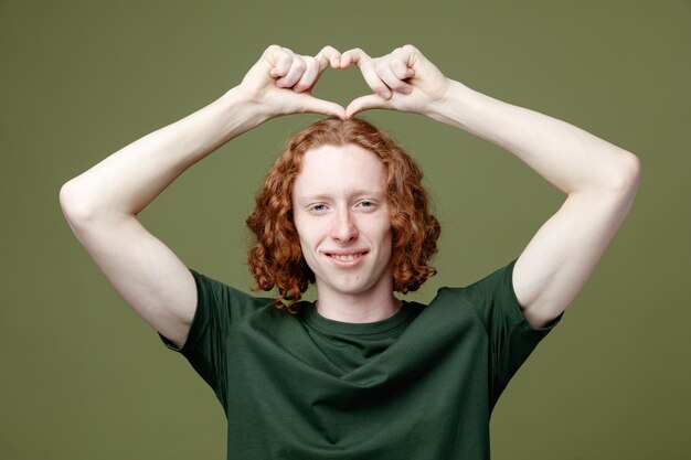 Улыбаясь, показывая жест сердца молодой красивый парень в зеленой футболке, изолированные на зеленом фоне