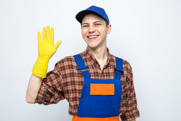 Smiling showing hello gesture young cleaning guy wearing uniform and cap with gloves 