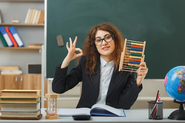 Sorridente che mostra gesto ok giovane insegnante femminile che indossa occhiali con in mano abaco seduto alla scrivania con strumenti scolastici in classe
