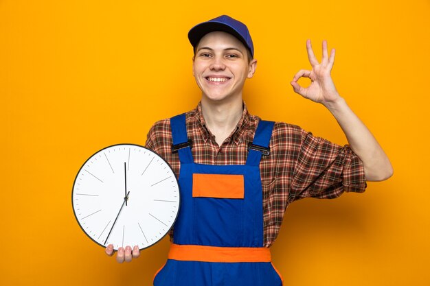 Smiling showing okay gesture young cleaning guy wearing uniform and cap holding wall clock 