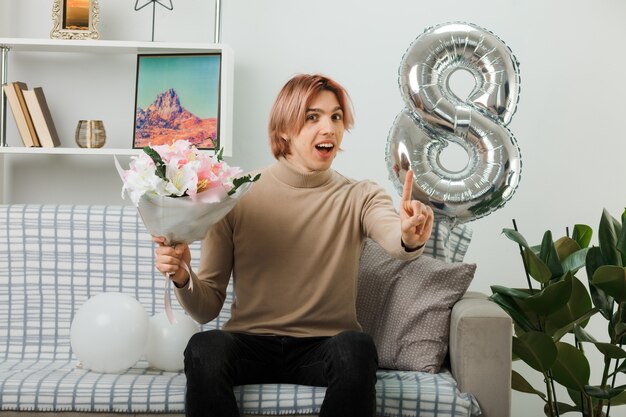 Smiling showing one handsome guy on happy women day holding bouquet sitting on sofa in living room