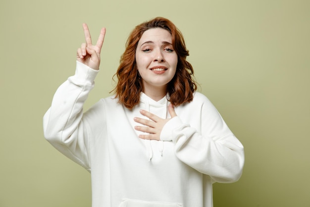 Smiling showing peace gesture putting hand on heart oung female wearing white sweater isolated on green background