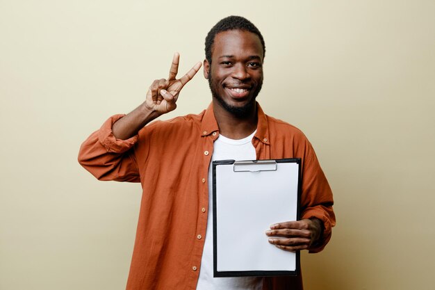 白い背景で隔離のクリップボードを保持している平和ジェスチャー若いアフリカ系アメリカ人男性を示す笑顔