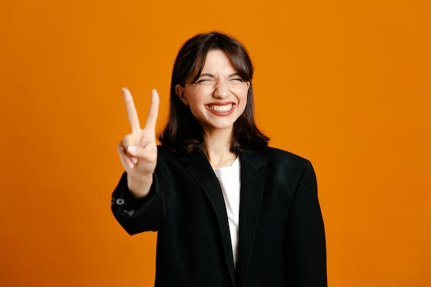 Улыбаясь, показывая мирный жест молодая красивая женщина в черной куртке на оранжевом фоне