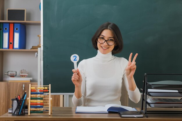улыбаясь, показывая мирный жест молодая учительница в очках, держащая номер вентилятора, сидящая за партой со школьными инструментами в классе