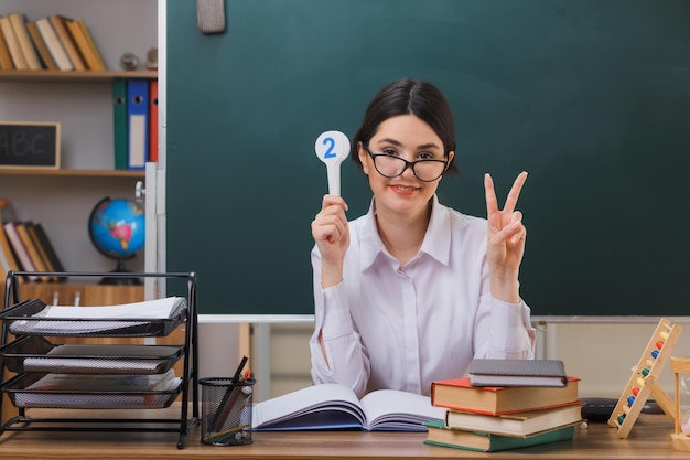 교실에서 학교 도구와 함께 책상에 앉아 번호 재미를 들고 안경을 쓰고 미소를 보여주는 평화 제스처 젊은 여성 교사