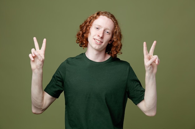 Улыбаясь, показывая мирный жест молодой красивый парень в зеленой футболке на зеленом фоне