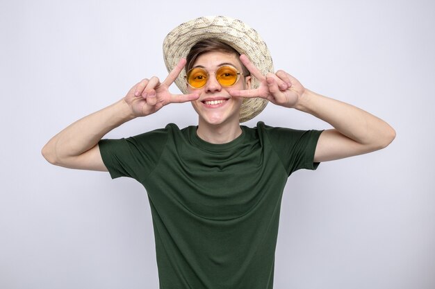 Smiling showing peace gesture young handsome guy wearing hat with glasses isolated on white wall