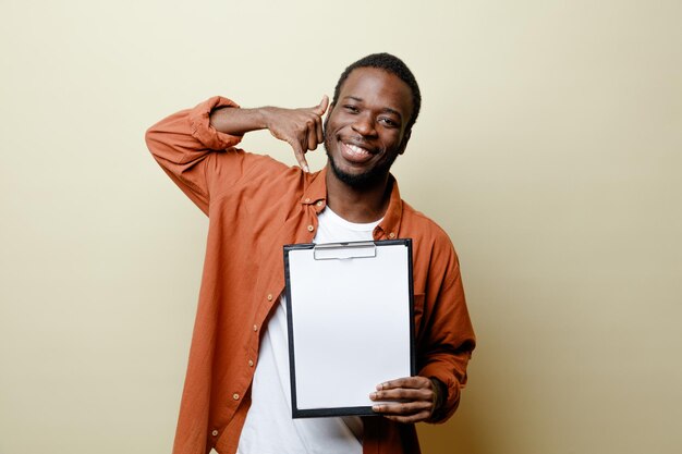 白い背景に分離されたクリップボードを保持している若いアフリカ系アメリカ人男性の電話ジェスチャーを示す笑顔