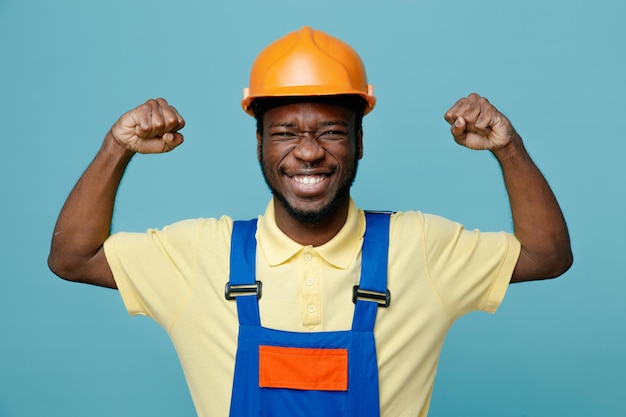 Foto gratuita sorridente che mostra gesto forte giovane costruttore afroamericano in uniforme isolato su sfondo blu