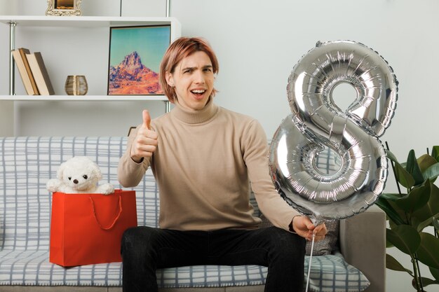Smiling showing thumb up handsome guy on happy women day holding number eight balloon sitting on sofa in living room