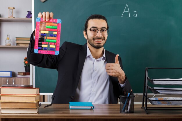 교실에서 학교 도구를 가지고 테이블에 앉아 있는 주판을 들고 안경을 쓴 남자 교사를 엄지손가락으로 보여주는 미소
