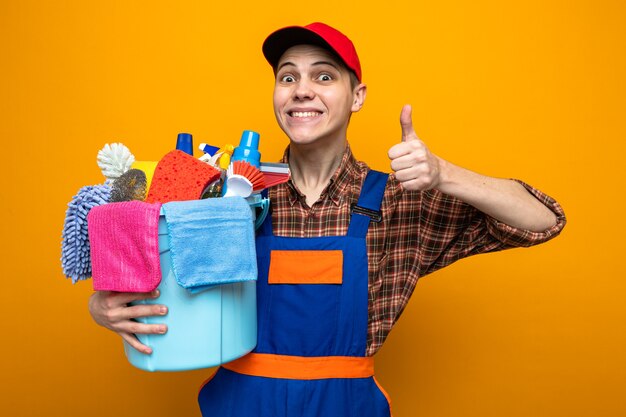 Smiling showing thumb up young cleaning guy wearing uniform and cap holding bucket of cleaning tools 