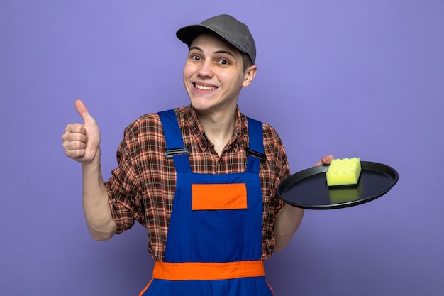 Foto gratuita sorridente che mostra il pollice in su giovane addetto alle pulizie che indossa l'uniforme e il cappuccio che tiene la spugna sul vassoio