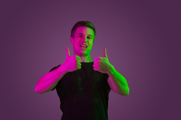 Улыбается, показывает палец вверх. Портрет кавказского человека на фиолетовом фоне студии в неоновом свете. Красивая мужская модель в черной рубашке. Понятие человеческих эмоций, выражения лица, продаж, рекламы.