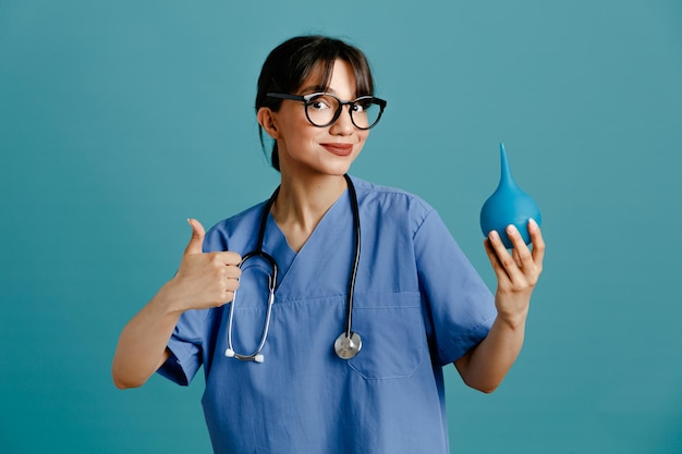 улыбаясь, показывая большие пальцы вверх, держа клизмы молодая женщина-врач в униформе фит стетоскоп, изолированные на синем фоне