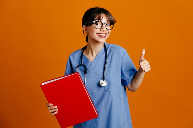 주황색 배경에 격리된 제복을 입은 젊은 여성 의사가 폴더를 들고 엄지손가락을 치켜들고 웃고 있습니다.