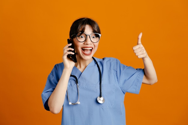 オレンジ色の背景に分離された均一なフィス聴診器を身に着けている若い女性医師が電話で話す笑顔