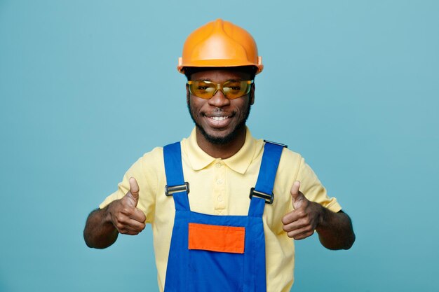 Smiling showing thumbs up young african american builder in uniform isolated on blue background