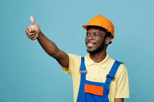 Sorridente mostrando pollice in alto giovane costruttore afroamericano in uniforme isolato su sfondo blu