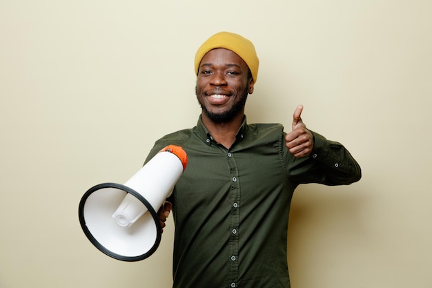 Smiling showing thumbs up young african american male in hat wearing green shirt holding loudspeaker isoloated on white background