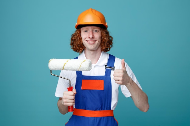 smiling showing thumbs up young builder man in uniform holding roller brush isolated on blue background