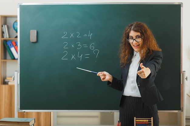 교실에서 포인터로 칠판에 칠판 포인트 앞에 서 있는 젊은 여성 교사 엄지손가락을 보여주는 미소