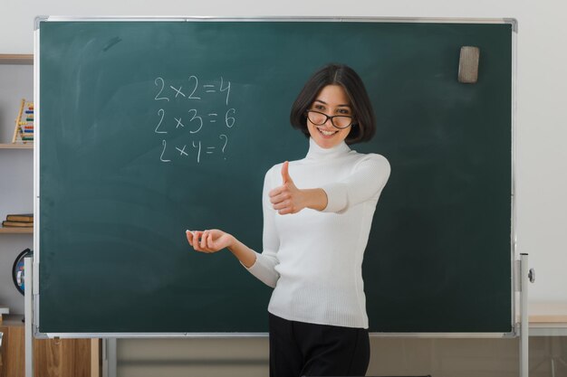 улыбаясь, показывая большой палец вверх, молодая учительница в очках стоит перед доской и пишет в классе