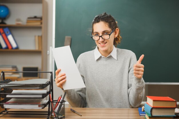 教室で学校の道具をつけて机に座って紙を持って眼鏡をかけている若い男性教師が親指を立てて笑っている