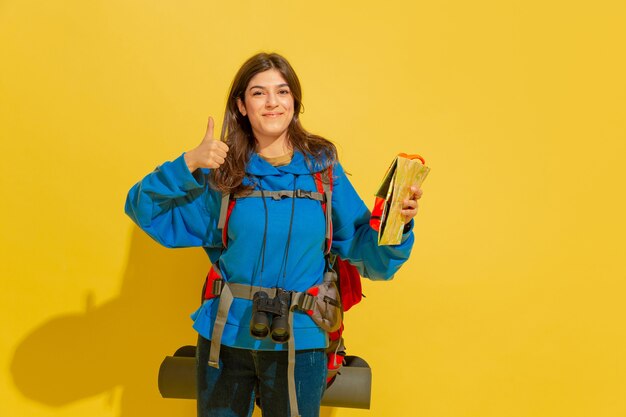 Smiling, shows thumb up. Portrait of a cheerful young caucasian tourist girl with bag and binoculars isolated on yellow studio background. Preparing for traveling. Resort, human emotions, vacation.