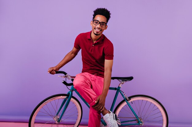 Улыбающийся застенчивый африканский парень в розовых штанах позирует с велосипедом. красивый темнокожий мужчина изолирован.
