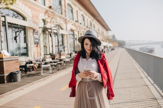 Smiling shy woman in hat standing on the street and looking at phone