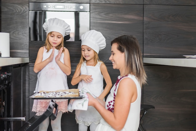 Free photo smiling siblings looking at mother holding fresh baked cookies in tray