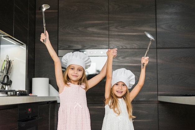 Smiling siblings with raised hands in kitchen holding ladle