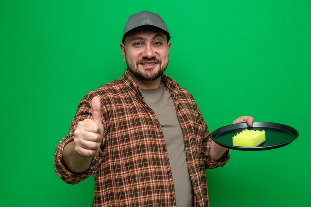 Smiling slavic cleaner man cleaning dishes with sponge and thumbing up 