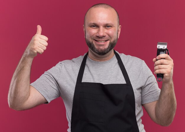 Smiling slavic middle-aged male barber in uniform holding hair clippers showing thumb up isolated on pink wall