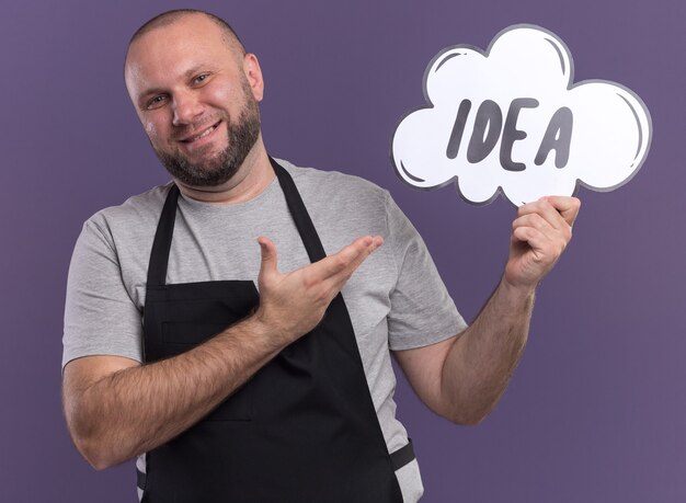 Smiling slavic middle-aged male barber in uniform holding and points at idea bubble isolated on purple wall