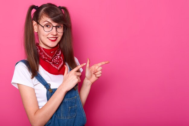 Smiling slim girl makes gesture with forefingers, showing the other way, looks pleased