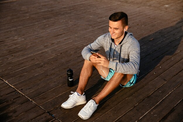 Smiling sportsman in earphones, listening to music, while using a mobile phone