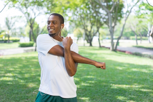 Smiling sporty black man listening to music and doing morning exercises in park.