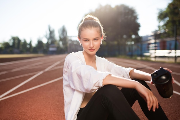 Sorridente ragazza sportiva in auricolari wireless che guarda premurosamente nella fotocamera mentre trascorre del tempo sulla pista da corsa dello stadio