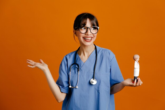 Smiling spreading hand holding toy young female doctor wearing uniform fith stethoscope isolated on orange background