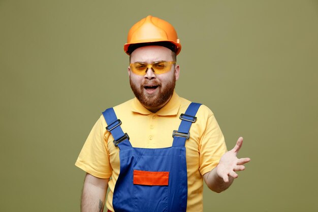 Smiling spreading hands young builder man in uniform isolated on green background