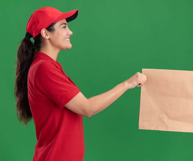 緑の壁に隔離されたクライアントに紙の食品パッケージを与える制服と帽子を身に着けたプロファイルビュー若い配達の女の子に笑顔
