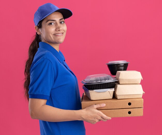 Smiling standing in profile view young delivery girl wearing uniform with cap holding food containers on pizza boxes isolated on pink wall