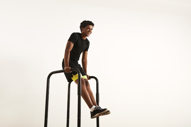 smiling strong African American athlete with an afro wearing black synthetic shirt and black and yellow shorts doing L-sits on short bars at home isolated on white.