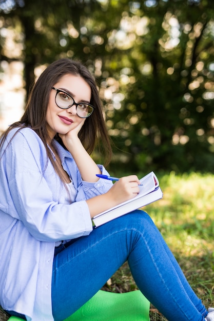 Smiling student girl in jeans jacket and glasses write in notebook in park