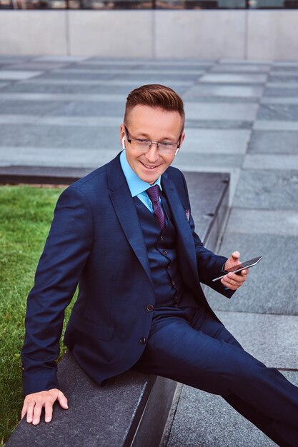 Free photo smiling stylish businessman dressed in an elegant suit holds a smartphone and looking away while sitting outdoors against skyscraper background.