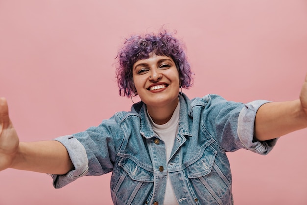 Sorridente signora elegante con i capelli corti ricci lilla in maglietta bianca e giacca blu prende selfie e ride.