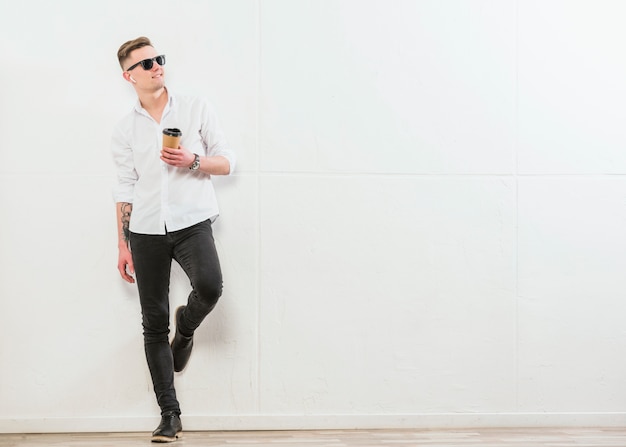 Усмехаясь стильный молодой человек держа на вынос устранимую кофейную чашку стоя против белой стены